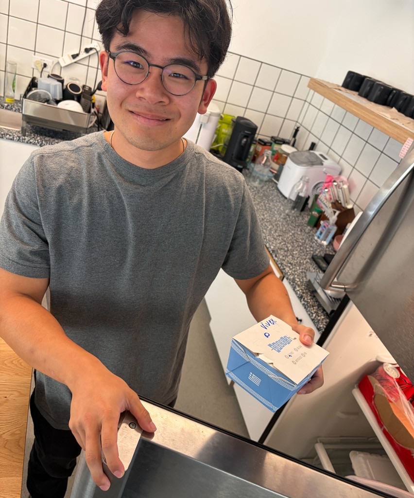 A teammate in a kitchen smiling while holding a takeout box by the fridge