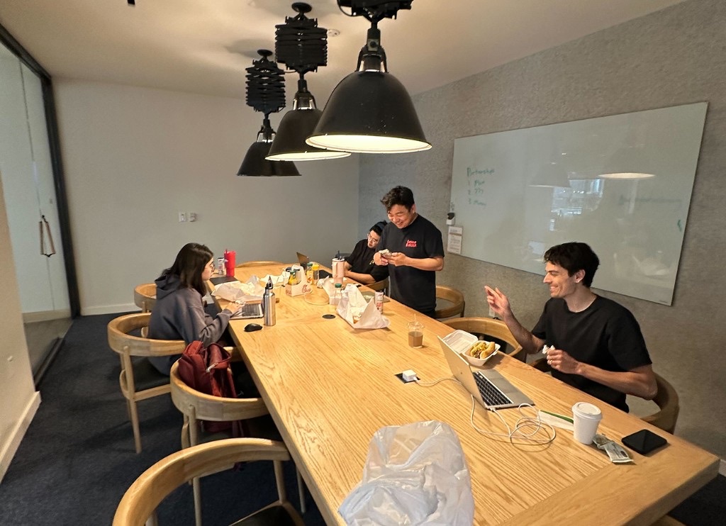 Teammates eating takeout around a long table under big pendant lights beside a whiteboard