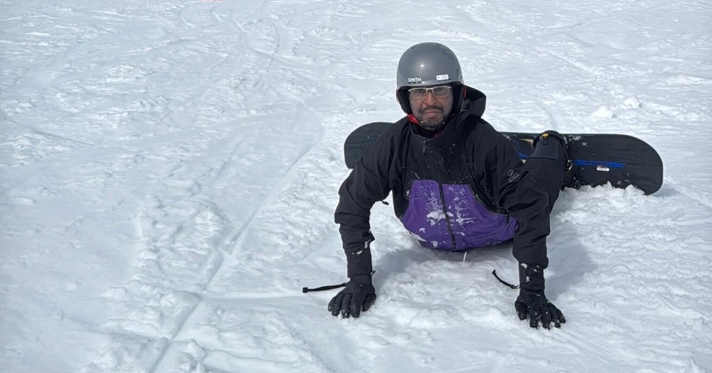A snowboarder lying face-down in the snow, grinning after a wipeout