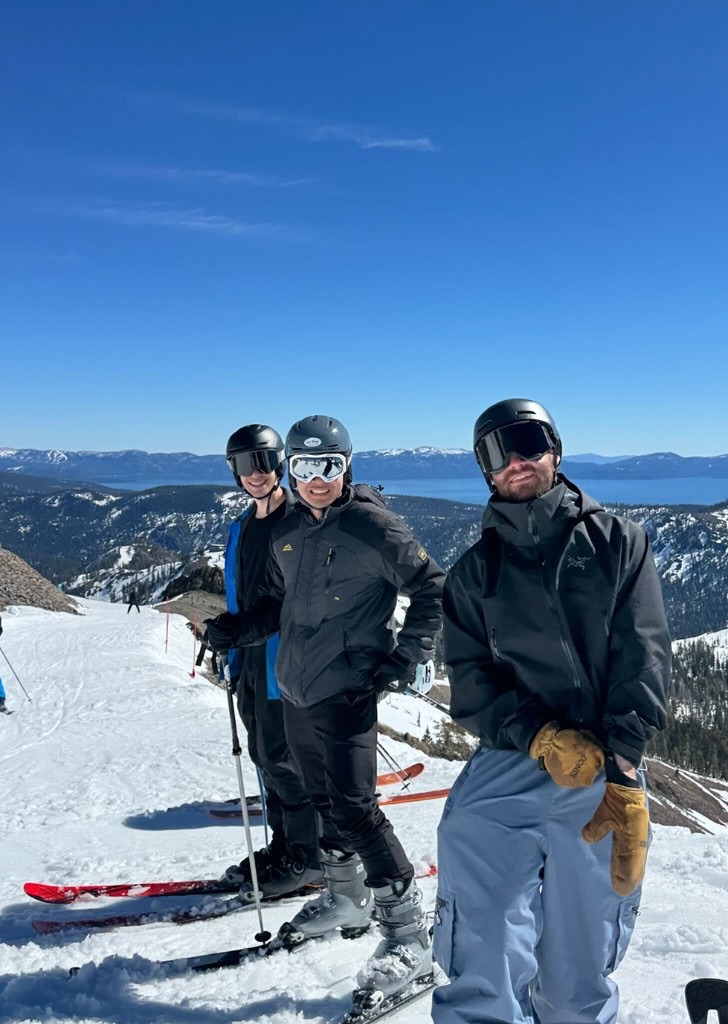 Three teammates in ski gear posing at the summit with Lake Tahoe in the distance